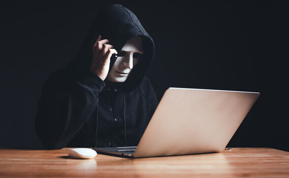 Black hat hacker in hood using laptop computer and call smartphone to victim on desk hacking privacy sensitive data hack in dark room background. Cyber security cyber crime concept. Hacking phishing