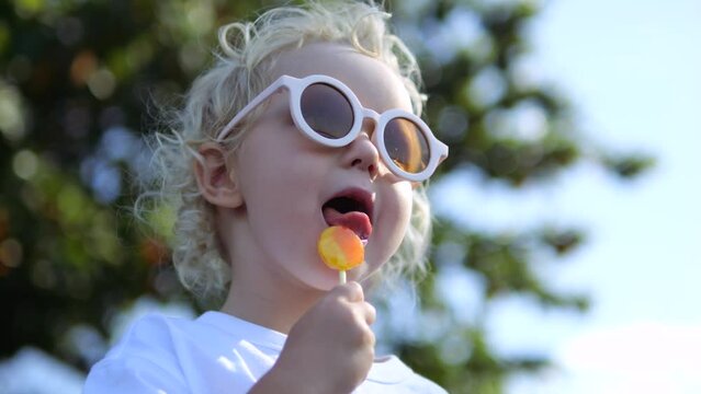 Close-up, low angle, slow motion, Caucasian blonde girl eating delicious sweet caramel lollipop. Little girl in round glasses enjoys taste of lollipop on street. Face of little girl eating lollipop
