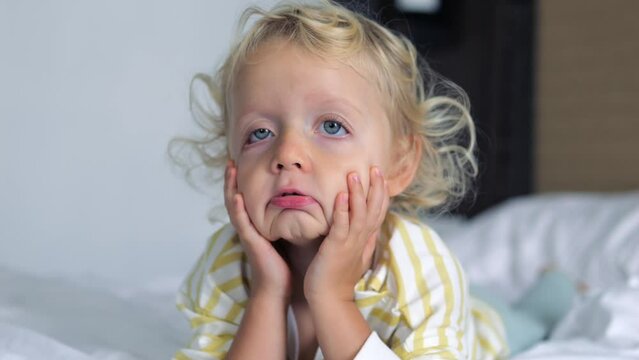 Cute funny child kid baby girl lying on bed and making faces, pulling skin of her face down, making faces and fooling around, having fun. Caucasian baby girl grimaces pulls skin of face down, close-up