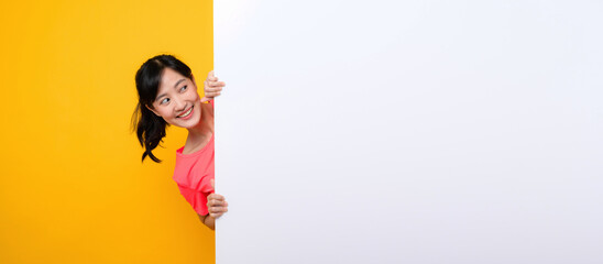young asian sports fitness woman happy smile wearing pink sportswear standing behind the white blank banner or empty space advertisement board against yellow background. wellbeing lifestyle concept. © Jirawatfoto