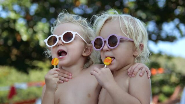 Slow motion close-up naked kids sisters in park on picnic outside licking lollipops on stick. Concept of happy childhood with variety of taste impressions. Naked kids eating lollipops slow motion