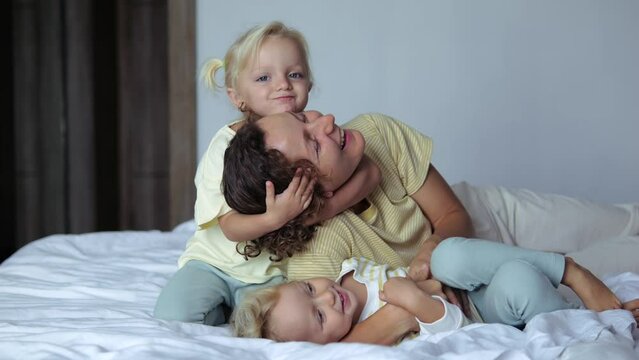Slow motion mom and kids having fun on bed hugging laughing smiling embracing loving each other spending time playing together. Two girls daughters hug their mom lying on bed, slow motion.