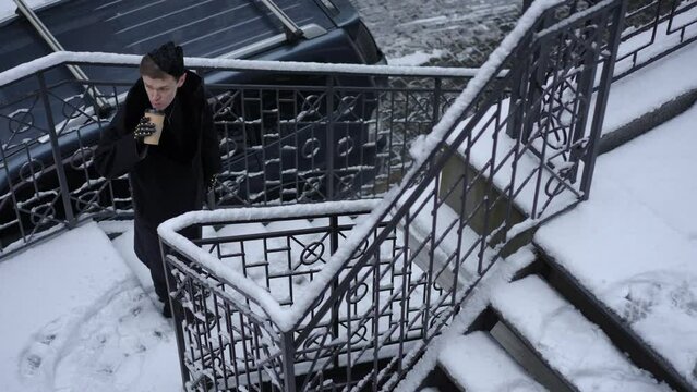 Wide shot high angle view LGBT person drinking coffee walking downstairs in cold winter urban city. Confident stylish Caucasian gay man with paper cup strolling in slow motion outdoors