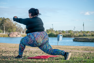 Young fat woman exercising yoga in the park by the lake. Healthy lifestyle