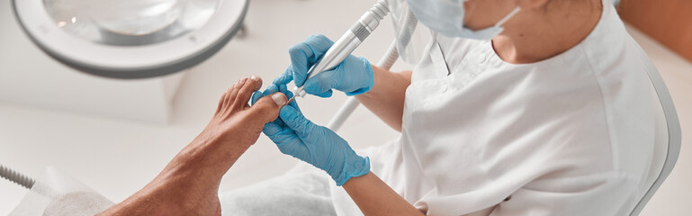 Lady uses grinding machine to polish toenail of male client at in beauty salon