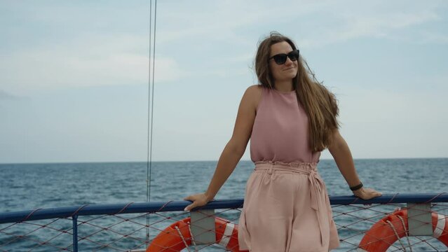 The young woman in pink shorts and a blouse stands on the upper deck of a cruise ship, smiling. slow motion