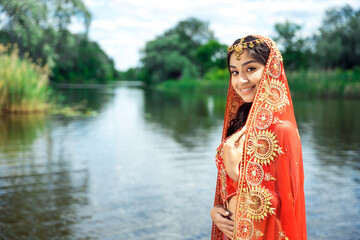 Fototapeta Beautiful Indian woman wearing traditional red dress posing against river. Bollywood style background with copy space. obraz