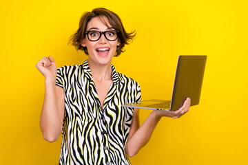 Portrait of ecstatic overjoyed woman wear striped blouse rising fist up hold laptop win gambling isolated on yellow color background