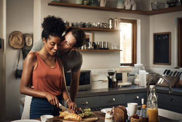 Interracial couple, kiss and cooking in kitchen for morning breakfast, love or caring relationship at home. Man kissing woman while making food, romantic meal or cutting ingredients on table in house - obrazy, fototapety, plakaty