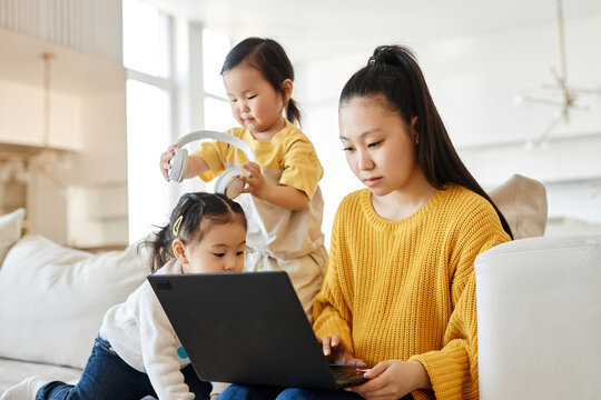 Asian young mom working on laptop while her children playing near her on sofa in the living room