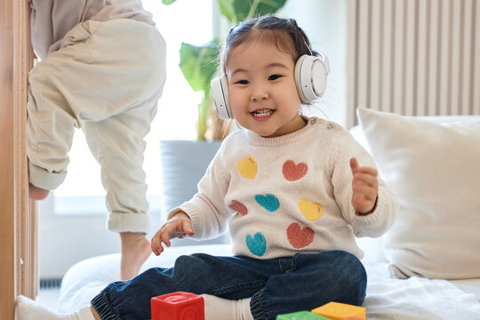 Little girl listening to music in wireless headphones while sitting on sofa with toys