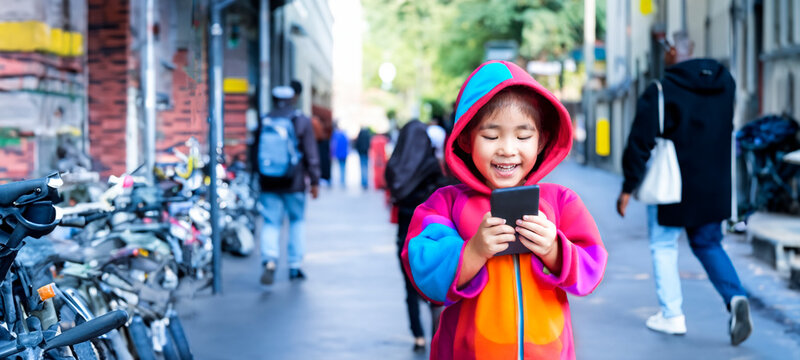 Asian Kids Using Smartphone In Coloring Hoodie Walking On Street. Mobile Addict Is New Behavior In Digital Generation.