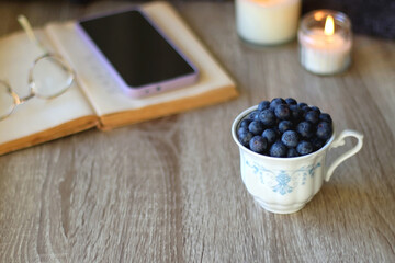 Cup of blueberries, set of books, phone, eyeglasses, lit candles and blanket on the table. Hygge at home. Selective focus.