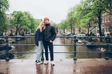 Smiling young couple on bridge over rainy canal in Amsterdam boats, trees, water, sweaters, hats, jeans, 20's