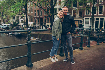 Smiling middle age couple on bridge over rainy canal in Amsterdam boats, trees, water, sweaters,...