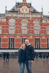 Smiling middle aged couple on in front of the red brick train station in Amsterdam in the rain wearing sweaters, hats, jeans, 40's