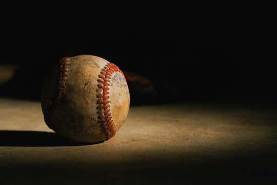 Low key lighting shows dark black shadows with vintage used baseball ball in spotlight for antique style sports art decor.