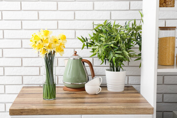 Fototapeta na wymiar Vase with blooming narcissus flowers, electric kettle, cups and houseplant on kitchen counter