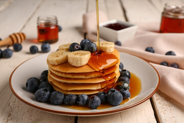 Pouring honey onto tasty pancakes with banana and blueberry on light wooden background, closeup - Powered by Adobe