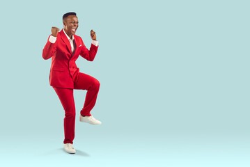 Fototapeta na wymiar Happy confident attractive African American man in cool modern trendy red suit celebrating victory, feeling overjoyed and super excited, fist pumping and having fun on light blue copyspace background
