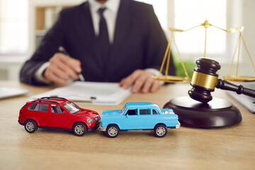 Close up of two toy model cars on background of judge who is conducting lawsuit in car accident. Toy cars representing accident stand on table next to judge's gavel the scales of justice.