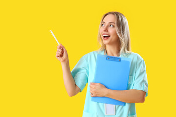Female medical intern with clipboard pointing at something on yellow background - obrazy, fototapety, plakaty