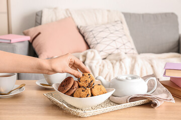 Woman holding cookie and teapot with cup of tea on wooden table in living room