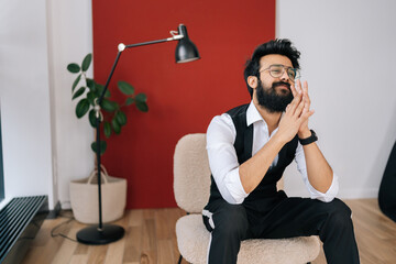 Medium shot of confident Indian businessman wearing elegant formalwear sitting on chair, smiling looking away. Front view of friendly Indian young man in fashion glasses posing at studio.