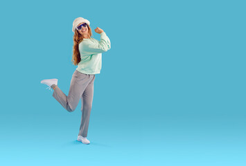 Full length portrait of happy laughing woman in white fluffy hat and sunglasses, mint sweatshirt and grey pants dancing on blue background with copy space standing on one leg. Advertisement banner.