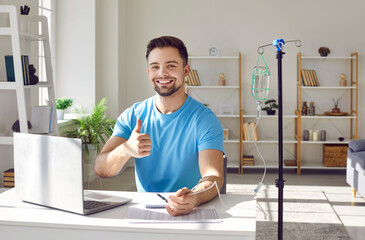Happy smiling man sitting and working on a laptop showing thumb up sign while receiving IV drip...