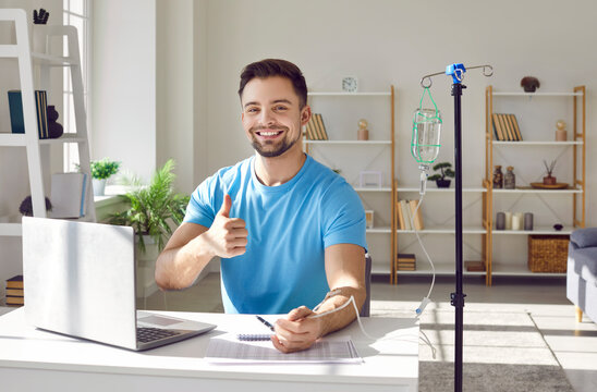 Happy smiling man sitting and working on a laptop showing thumb up sign while receiving IV drip infusion and vitamin therapy in his blood at the desk. Male person receiving injection therapy.