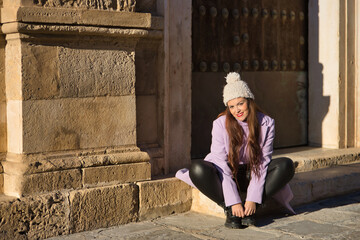 Young woman, beautiful, straight brown hair, leather pants, sweater, coat and wool hat, sitting on...