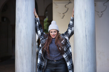 Young woman, beautiful, straight brown hair, leather pants, sweater, coat and wool hat, leaning on marble columns, smiling and happy. Concept beauty, fashion, autumn, winter, cold.