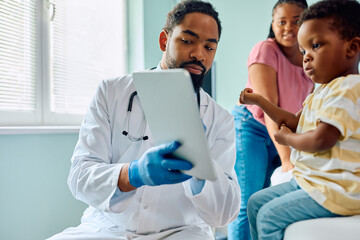 Black pediatrician and small boy using digital tablet at doctor's office.