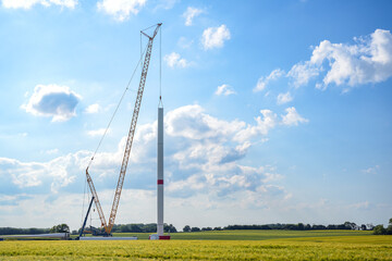 Fototapeta na wymiar Construction site of a wind turbine, tall crane installing a tube on the tower, nacelle, rotor hub and blades lying still on the ground, renewable energy and power concept, blue sky