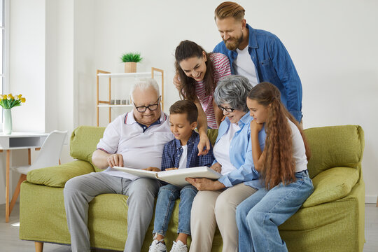 Family looks at photo album together and remembers stories from past during reunion of relatives. Elderly couple and their adult children and grandchildren are looking at photo while sitting on sofa.