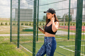 A young beautiful girl in sportswear and a cap inserts headphones into her ear and turns on music on her smartphone before running. Daily jogging in the fresh air