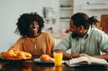 Fototapeta na wymiar A cheerful african american wife and husband sitting together at the breakfast table at home and smiling at each other.