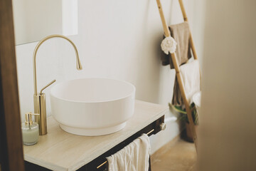 Modern ceramic sink with golden faucet on rustic stand with soap and towel on background of bathtub, modern bathroom interior. Stylish boho bathroom design.