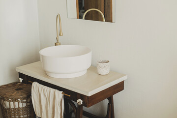 Modern ceramic sink with golden faucet on rustic stand with soap and towel on background of wall with mirror and travertine tile, modern bathroom interior. Stylish boho bathroom design.