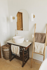 Modern bathroom interior. Modern ceramic sink with golden faucet on rustic stand with soap and ladder with towel on background of wall with mirror. Stylish boho bathroom design.