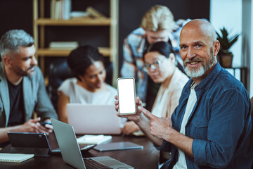 Mature man holds up mobile phone with white copy space on screen, offering blank canvas for ideas. In background, business team in loft office engages in brainstorming session, collaborating and