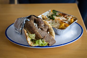 A Greek Mediterranean gyro wrapped in foil on a plate with a side of Greek fries covered in melted mozzarella and feta cheese, herbs, and spices