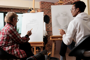 Diverse different generation people drawing on canvas and talking during creative art lesson in modern studio. Senior woman teaching African American student guy how to draw naturalistic still life