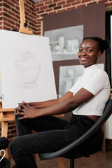 Fototapeta na wymiar Portrait of happy smiling African American girl sitting at easel feeling satisfied with work of art she created during drawing class. Young cheerful woman learning new things and having fun