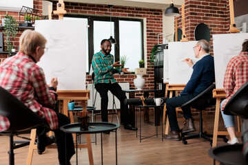 Friendly African American man teacher guiding group of different aged people through basics of drawing at art class. Young guy instructor explaining sketching techniques to students during workshop
