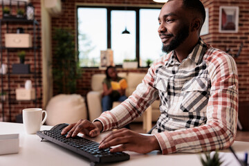 Obraz premium Smiling african american freelancer working remote typing and looking at computer screen while roommate is relaxing on couch. Male programmer using pc to develop software at home reading emails.