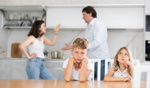 Confused, upset brother and sister are sitting at home during mom and dad quarrel. Children suffer because of their parents discord. Husband and wife screaming at each other in background blurred