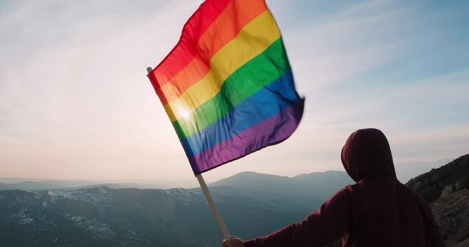 LGBT flag on sunset background by the peak of mountain. LGBT gay pride flag, lesbian, gay, bisexual, transgender social movements. Concept of love same-sex couple. A flag waving against the wind