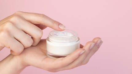 Beautiful graceful fingers of a woman scoop cream from a round glass jar on a pink background. Hand care concept. Layout for cosmetic products.